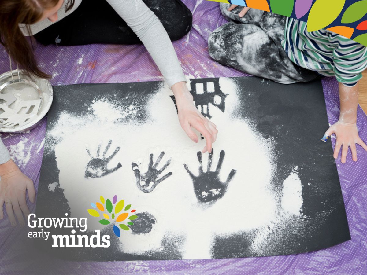 Parent and child on the ground with black cardboard and baby powder drawing shapes and hand prints
